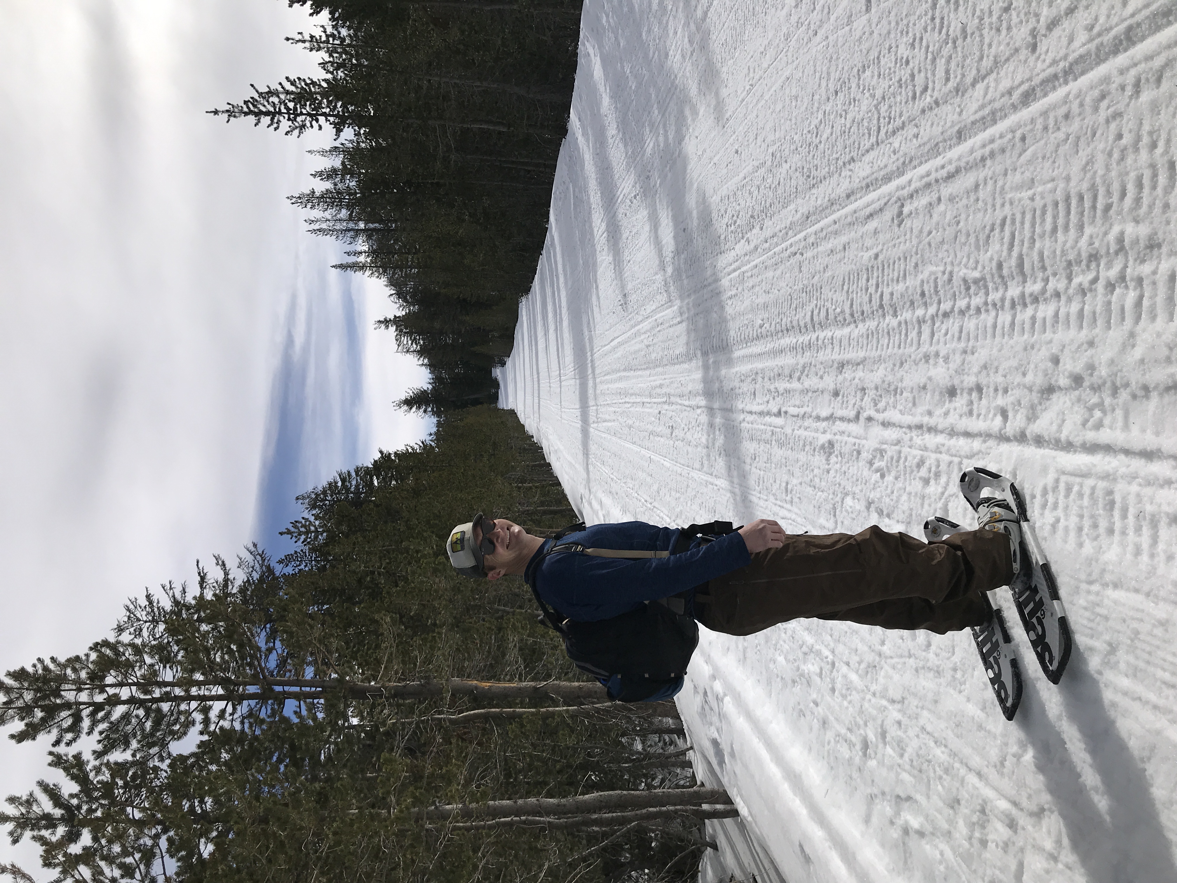 Man wearing sunglasses and a cap snowshoeing on a wide snow-covered trail lined with pine trees.