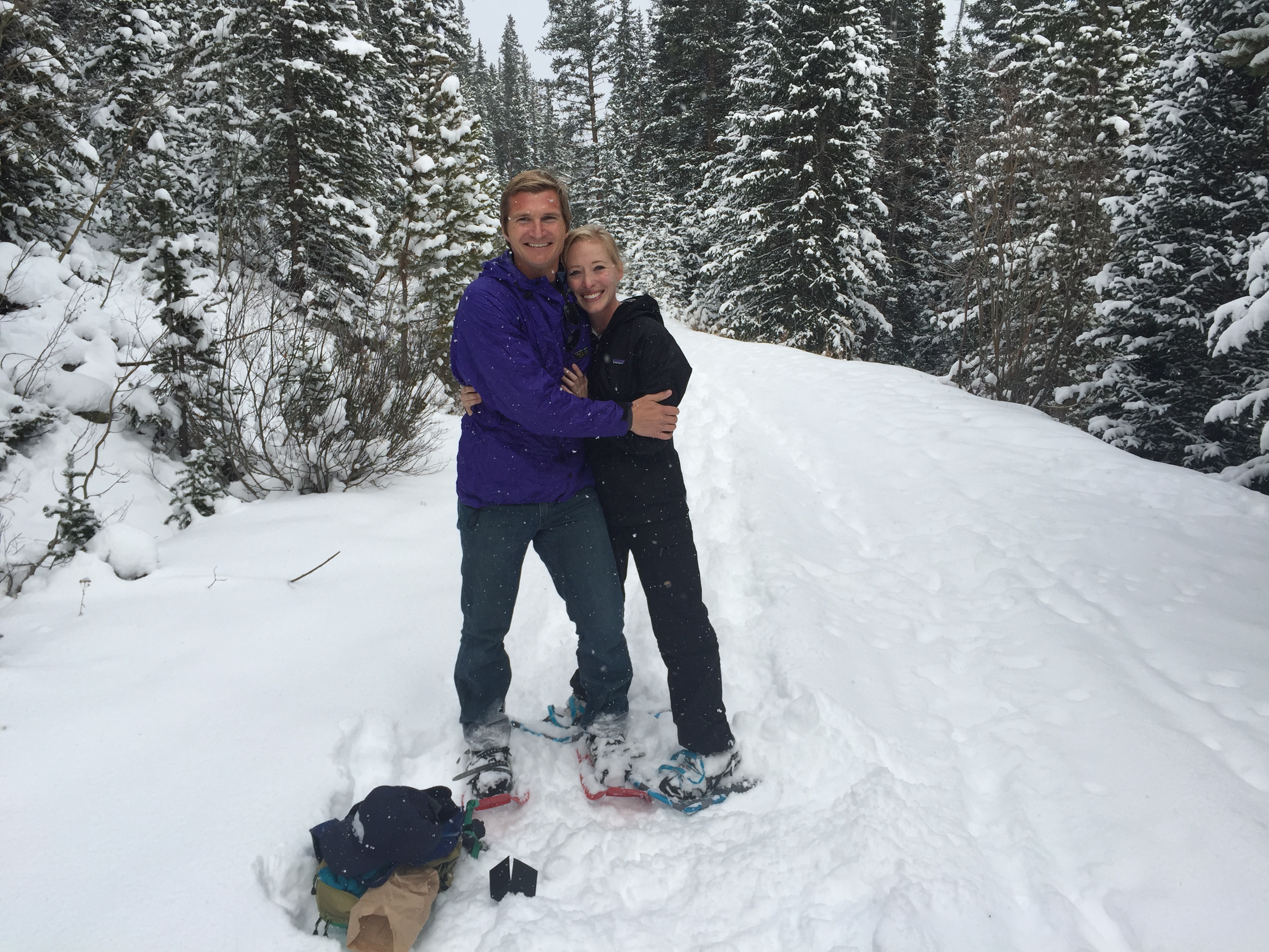 Couple smiling and hugging while standing on snowshoes on a snowy trail surrounded by snow-covered pine trees.