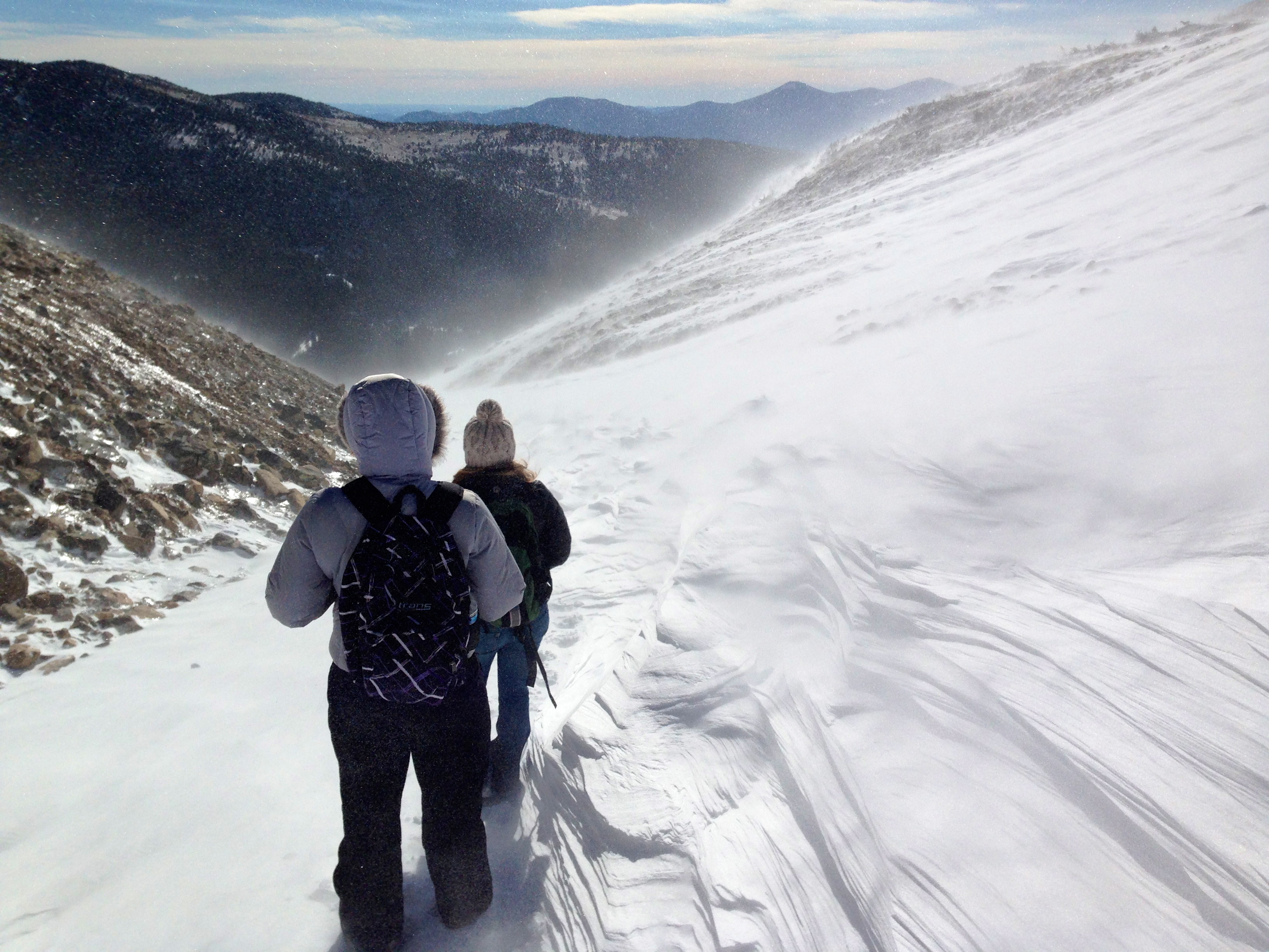 Two hikers wearing winter jackets and backpacks walking on a snowy mountain trail with wind blowing snow around.