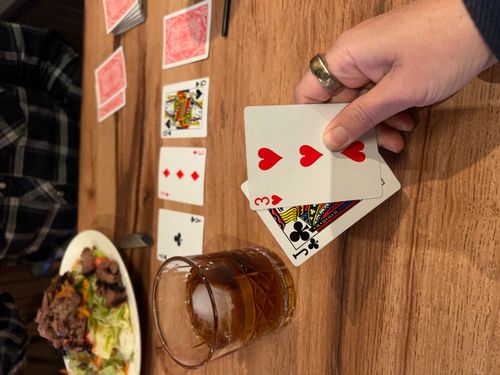 Hand holding two playing cards, three of hearts and jack of clubs, with a glass of brown drink and ice on a wooden table, food plate and person in background.