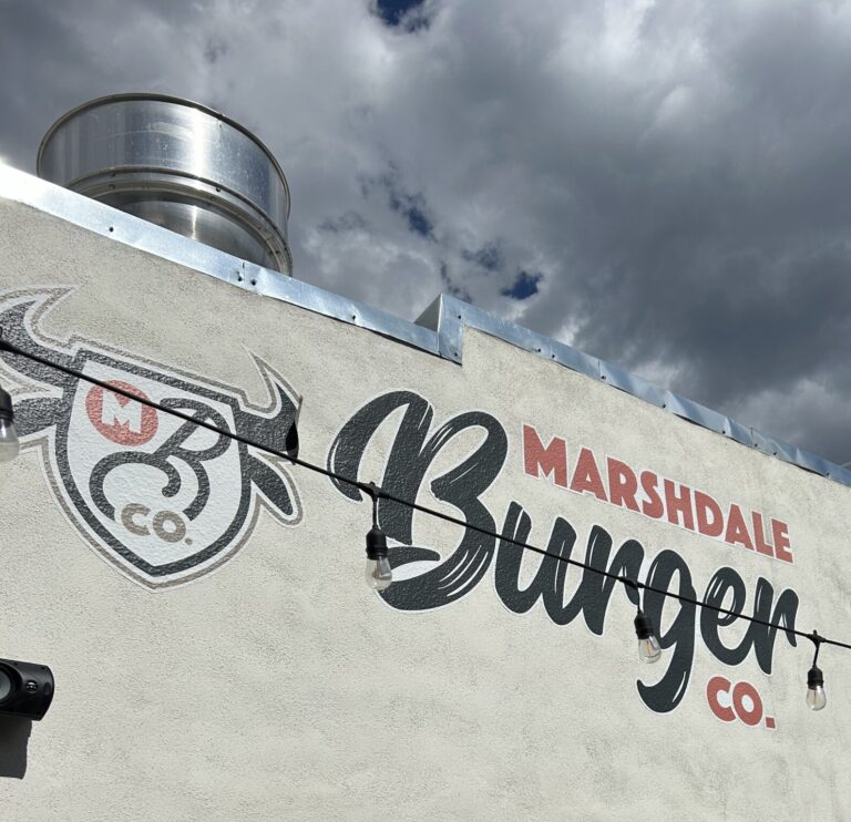 Exterior wall with Marshdale Burger Co. logo and lights hanging against a cloudy sky.