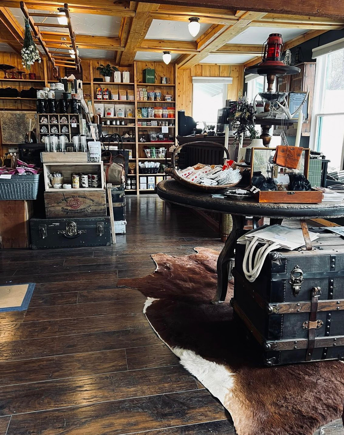 Rustic wooden store interior with shelves of packaged goods, a round table with baskets and boxes, and a cowhide rug on dark wooden floor at Blackfox Provisions, Evergreen.