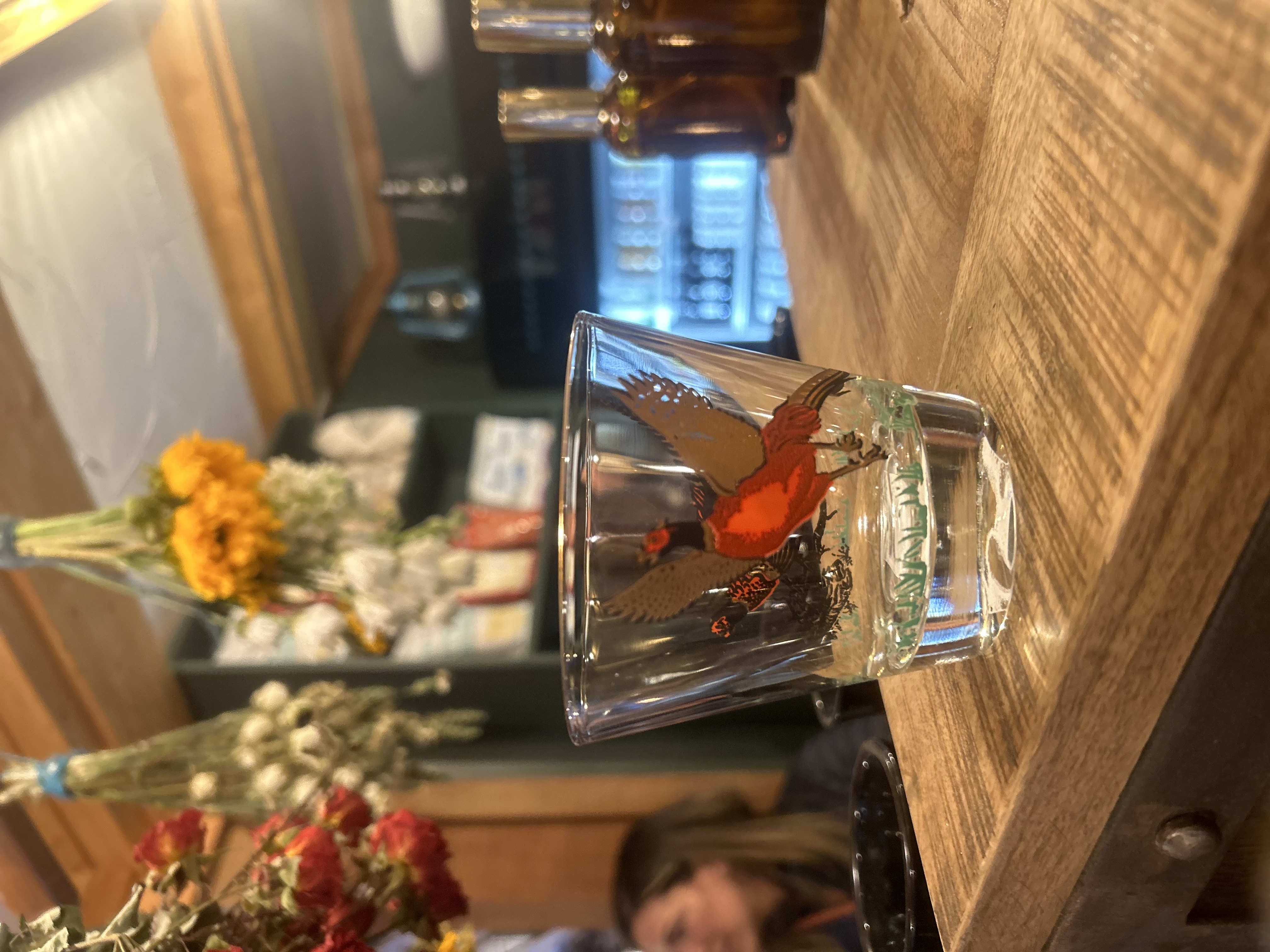 Clear glass with a colorful bird illustration placed on a wooden counter, with dried flowers hanging above and blurred background of a person and bottles at Blackfox Provisions, Evergreen.