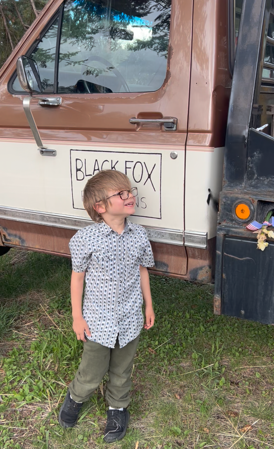 Young boy with glasses standing on grass beside a brown and beige truck with a sign reading 'BLACK FOX'.