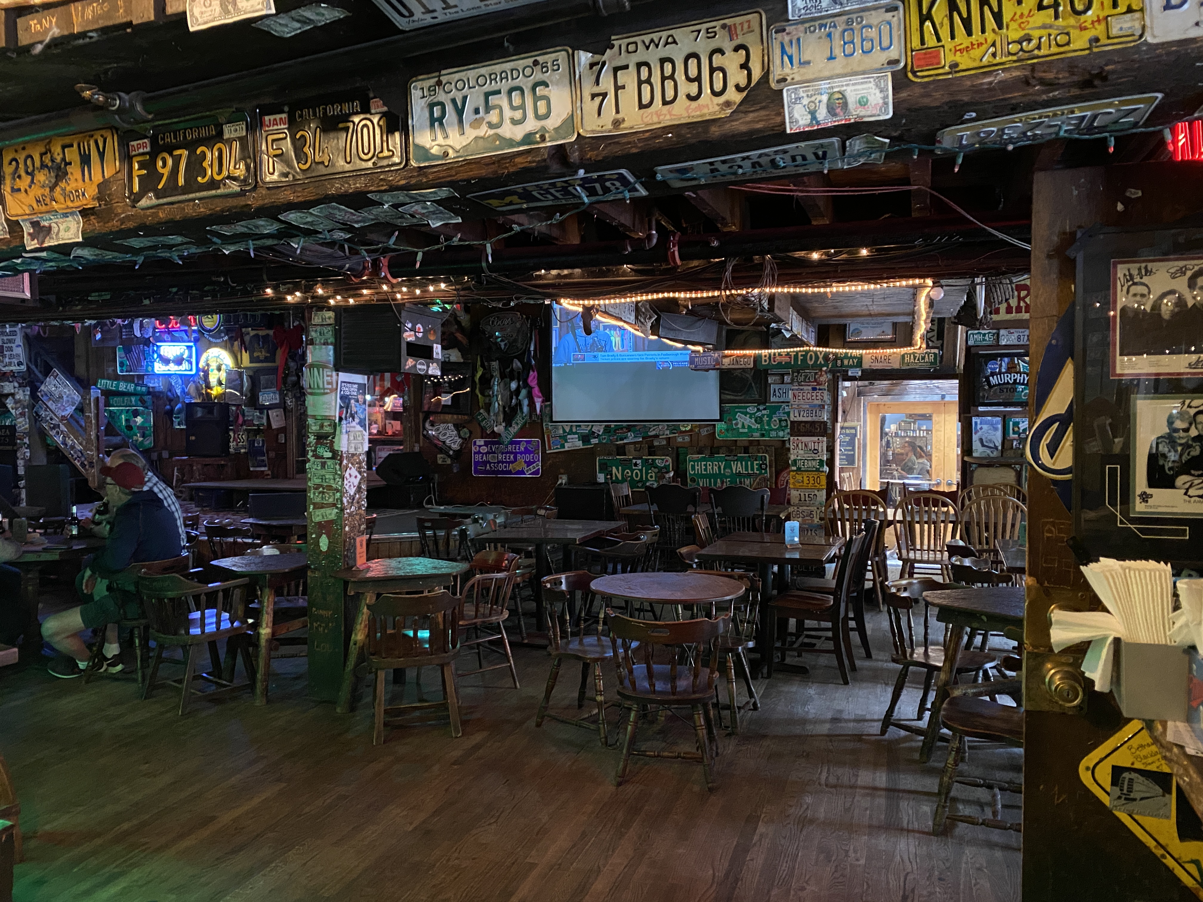 Interior of The Little Bear, a rustic bar with wooden floors, scattered tables and chairs, vintage license plates decorating the ceiling, and a few patrons seated.