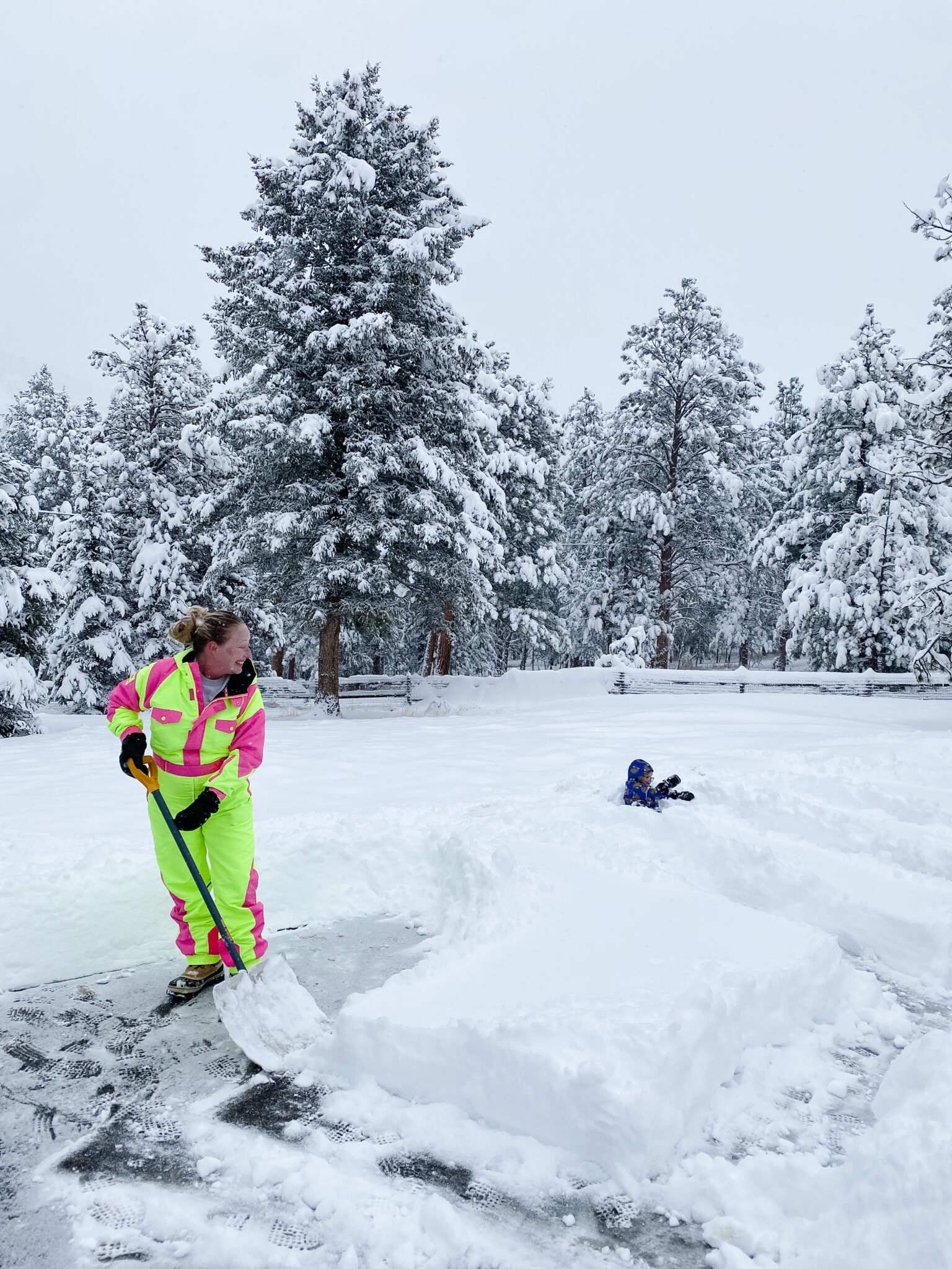 Person in bright yellow and pink snow gear shoveling snow on a driveway with a child playing in deep snow nearby, surrounded by snow-covered pine trees.