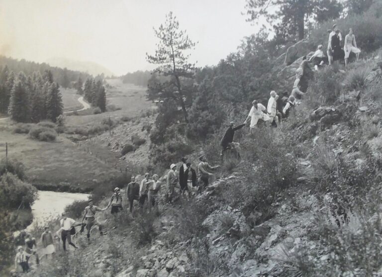 Black and white photo of a large group of people climbing a rocky hillside in a mountainous rural area with trees and a stream in the background in Evergreen, Colorado.