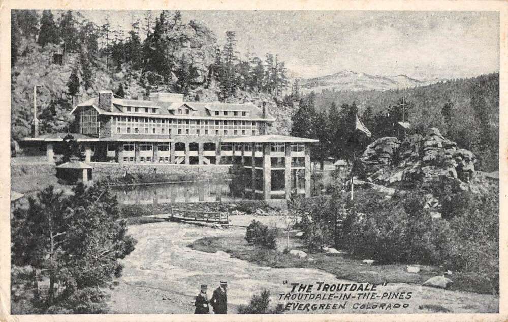 Black and white photo of the Troutdale hotel building nestled among pine trees with mountains in the background and two men walking on a path in the foreground.