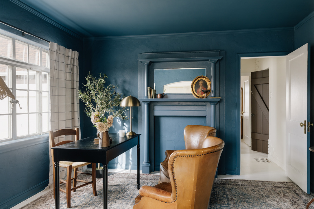 Cozy room with dark blue walls, a black desk with flowers and a lamp, a wooden chair, and a tan leather armchair near a decorative fireplace.