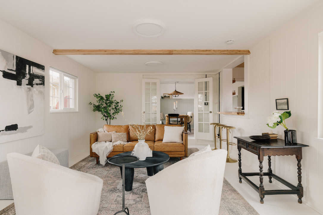 Bright living room with white walls, a brown leather sofa, two white armchairs, a black coffee table, and decorative plants and artwork.