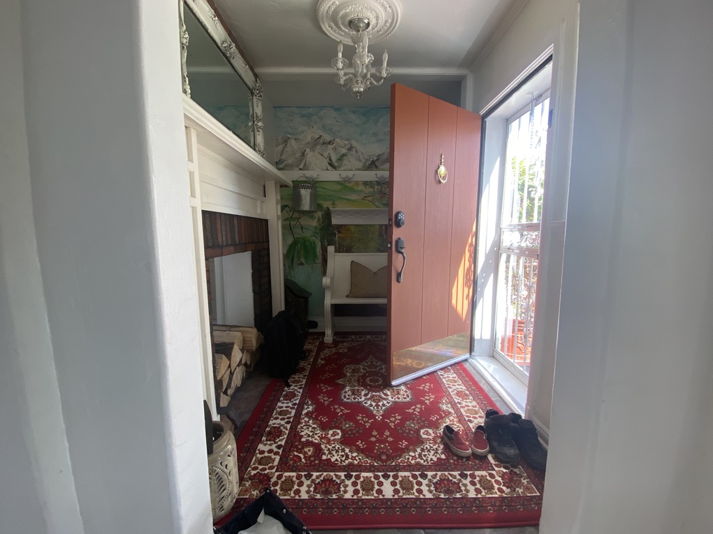 Entryway with open orange door, red patterned rug, white fireplace with stacked firewood, and bench with pillow under landscape mural.