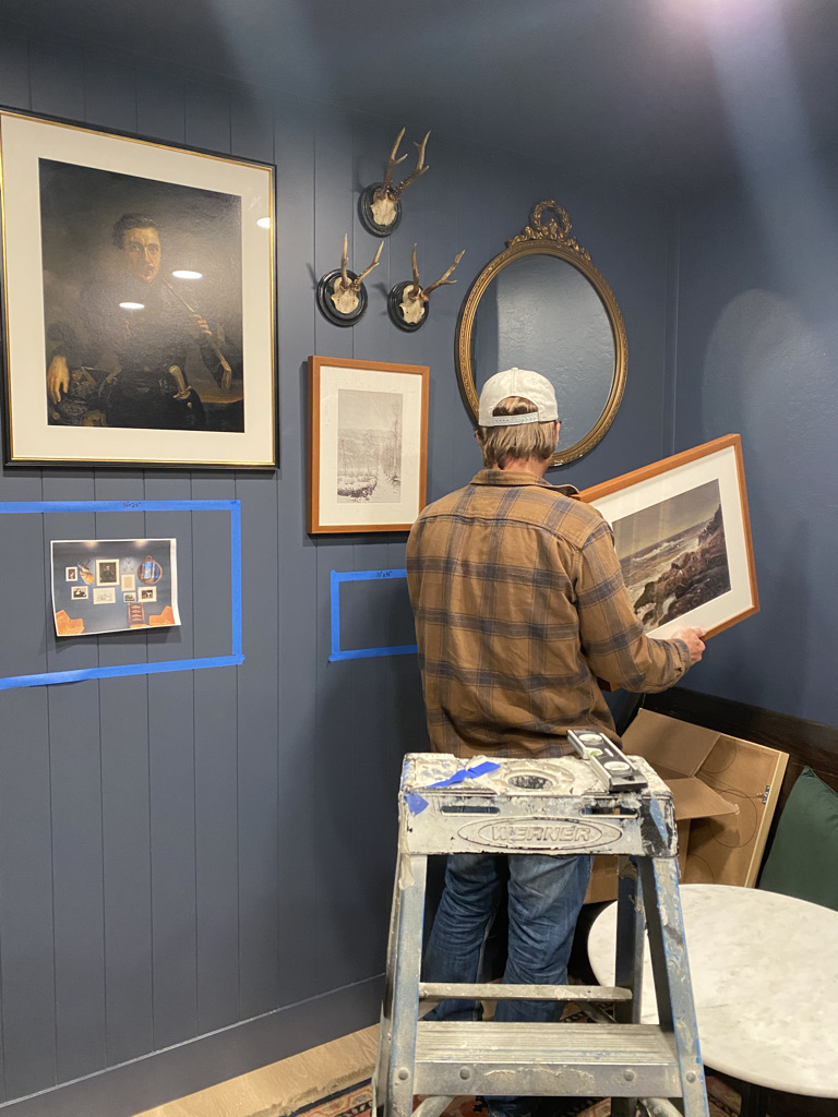 Man in a beige cap and plaid shirt hangs framed artwork on a blue wall with mounted antlers and a gold-framed oval mirror nearby.