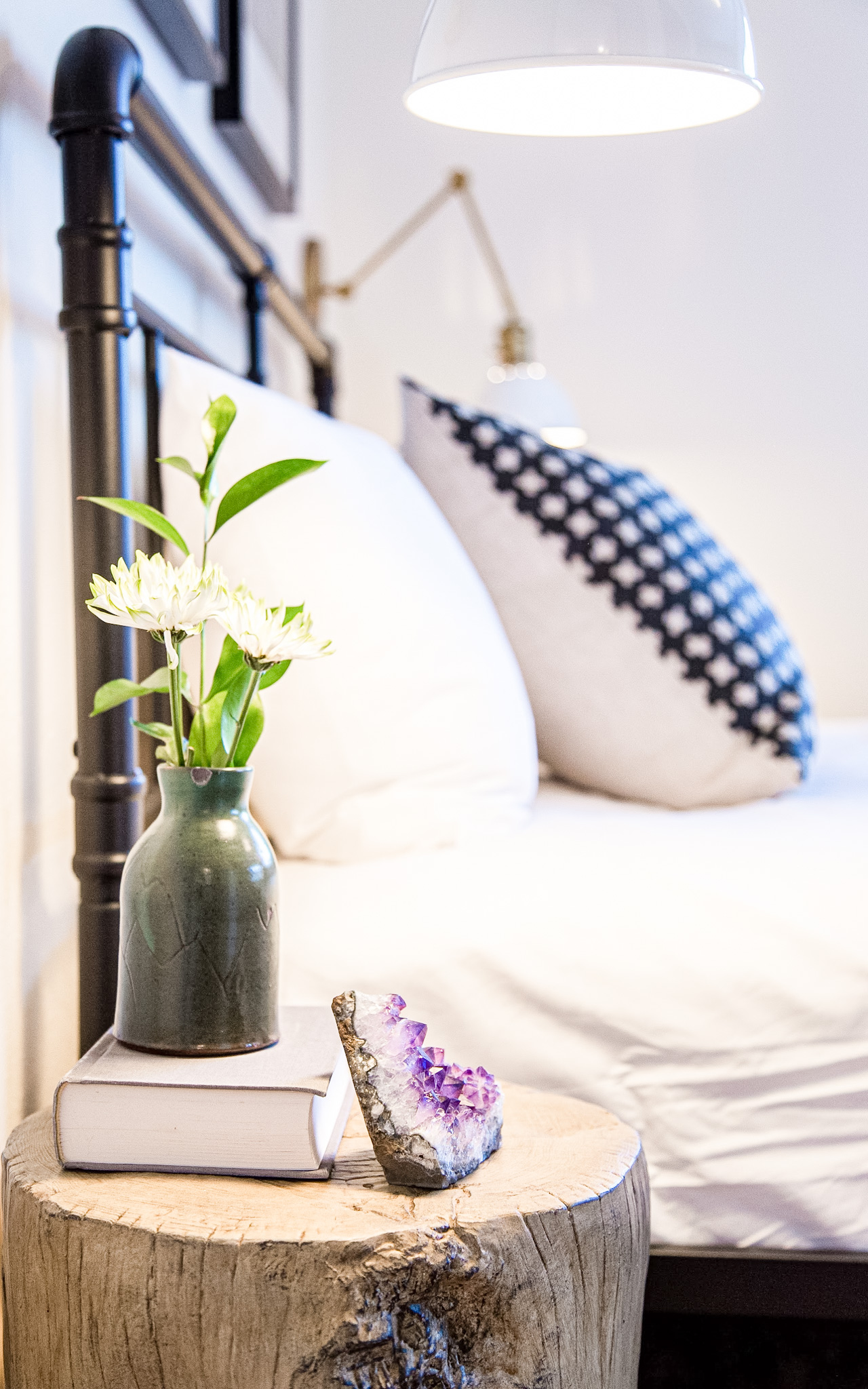 Rustic wooden stump side table with a gray vase holding white flowers, a closed book, and a purple amethyst crystal next to a bed with white and patterned pillows.