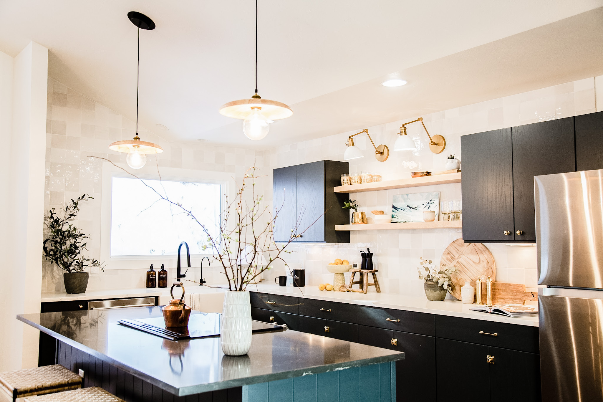 Bright modern kitchen with black cabinets, white tiled walls, stainless steel refrigerator, and a central island with a vase of branches and copper kettle.