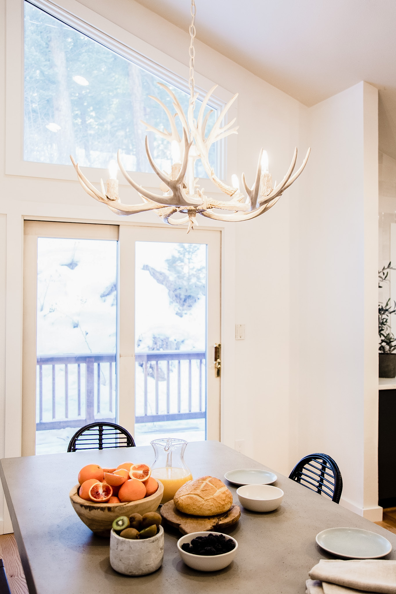 Dining table with a bowl of blood oranges, a loaf of bread, kiwi fruit, a pitcher of orange juice, and empty plates under a rustic antler chandelier near glass doors.