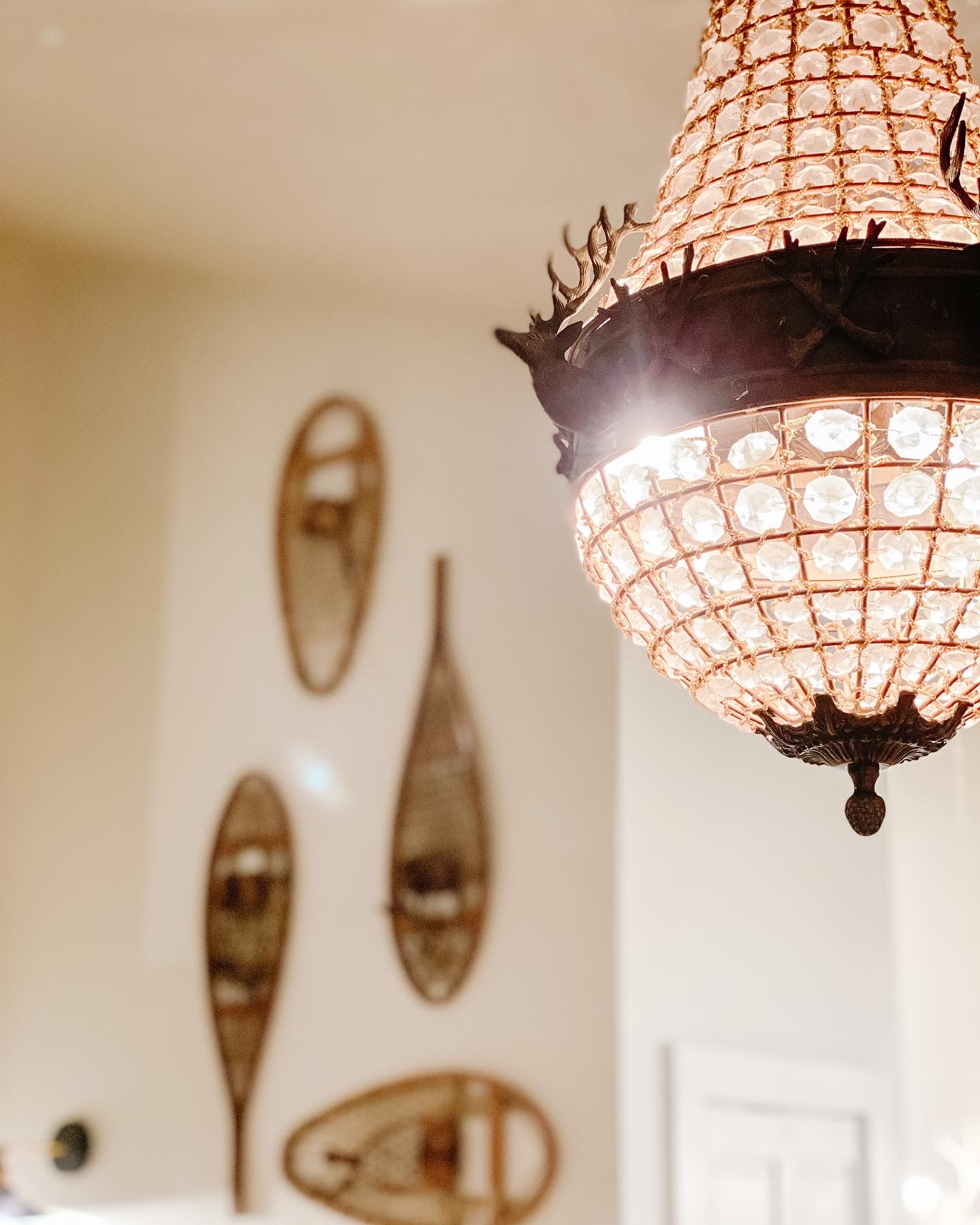 Crystal chandelier with decorative metal deer head accents hanging in a room with vintage snowshoes displayed on the wall.
