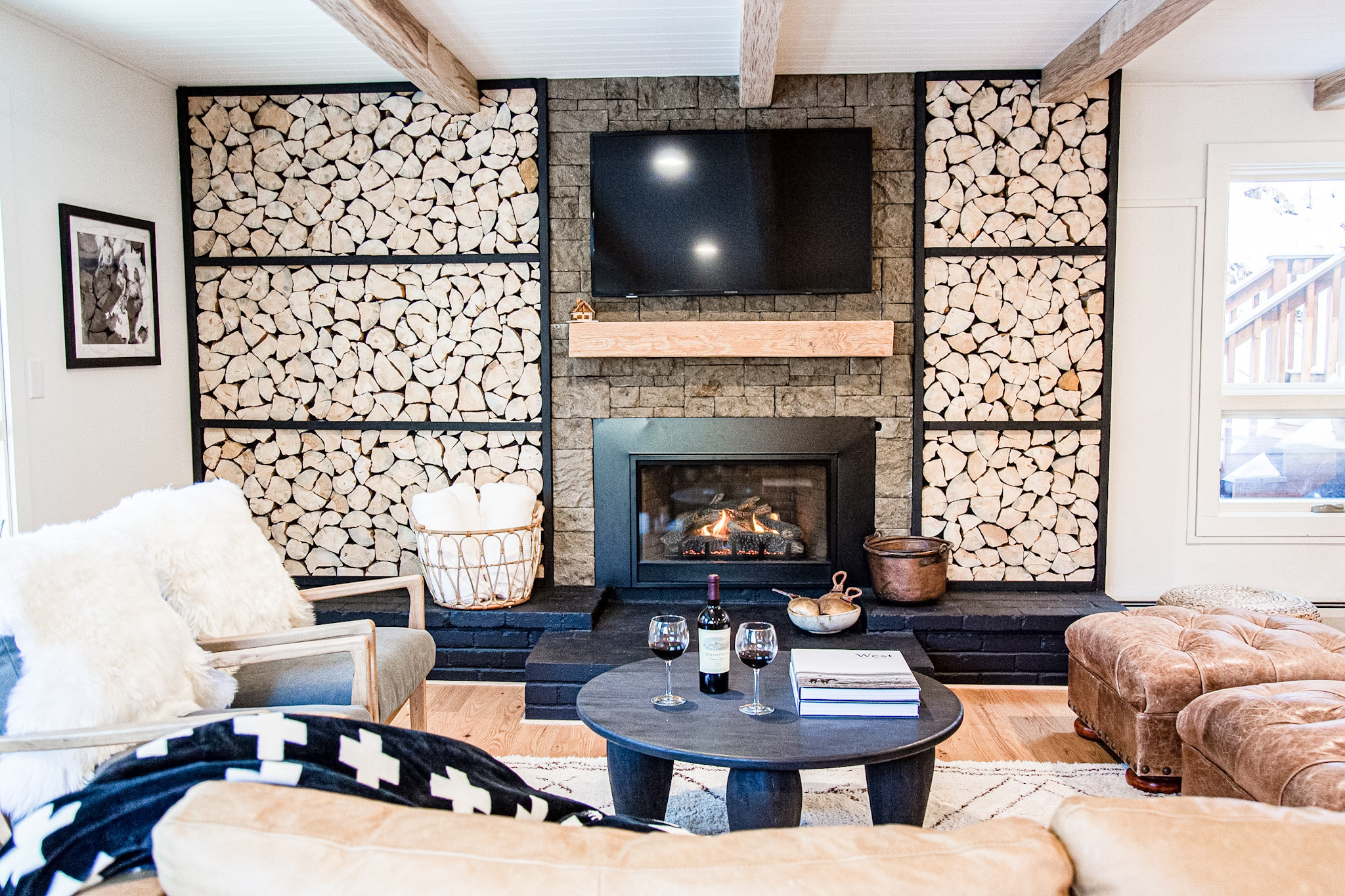 Cozy living room with stone fireplace, stacked firewood, a mounted TV, round coffee table with wine and books, and plush seating.