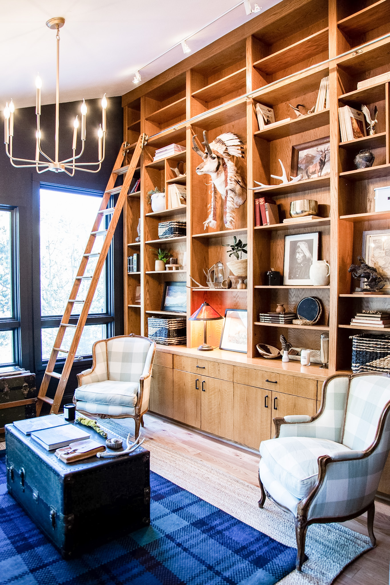 Cozy library corner with wooden floor-to-ceiling shelves, plaid armchairs, a wooden ladder, and a vintage trunk as a coffee table.