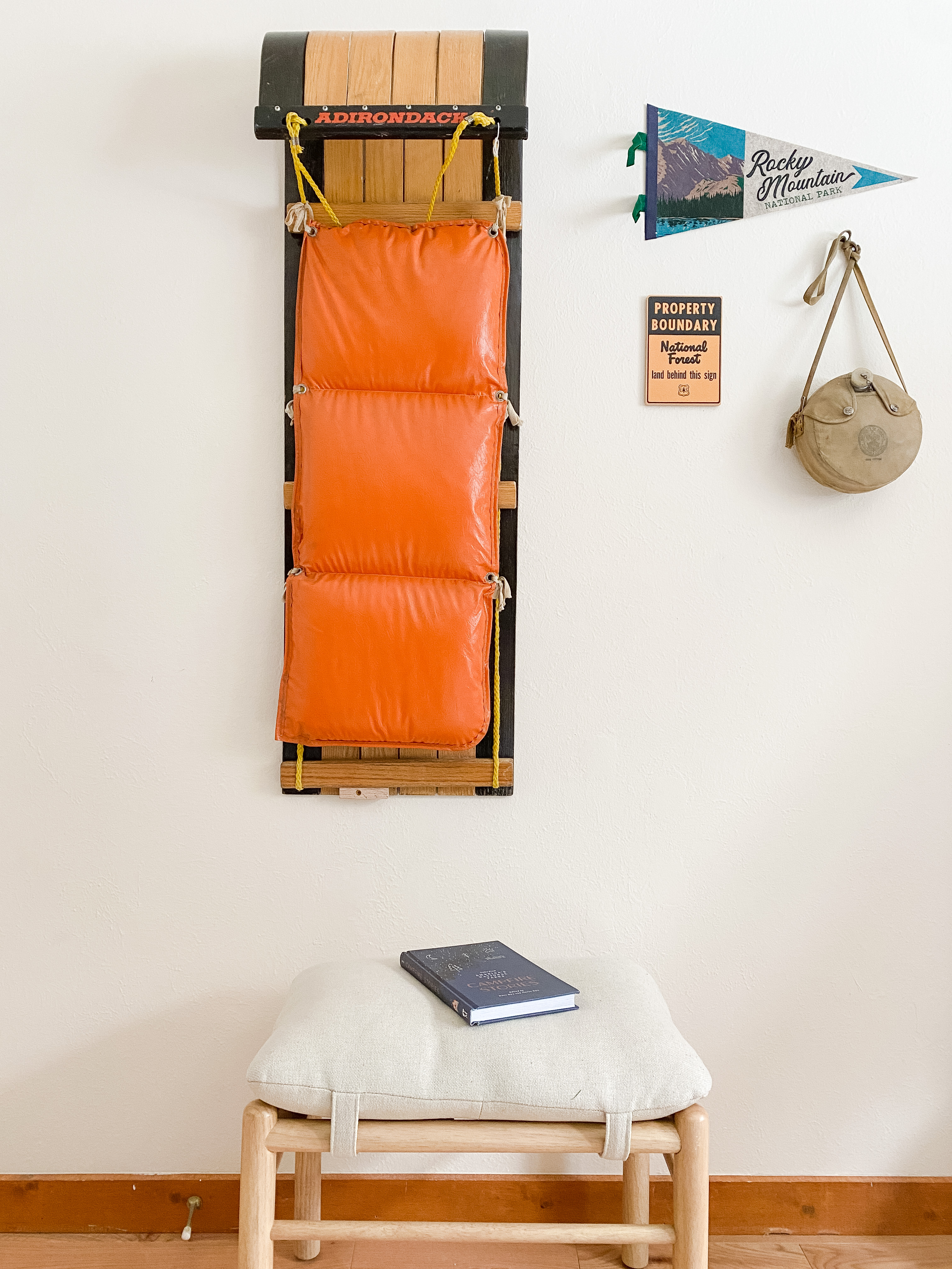 Orange Adirondack life jacket hanging on a wooden wall rack with a Rocky Mountain National Park pennant, a property boundary sign, and a vintage canteen beside it, above a cushioned wooden stool with a closed book.