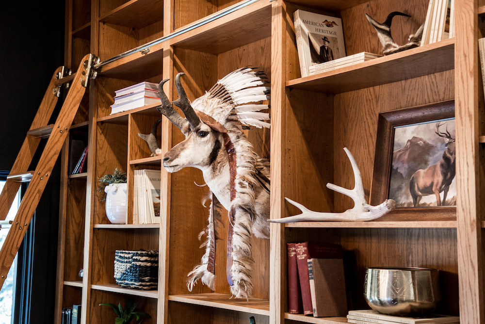 Wooden bookshelf with a mounted pronghorn wearing a feathered Native American headdress, books, antlers, and decorative items.
