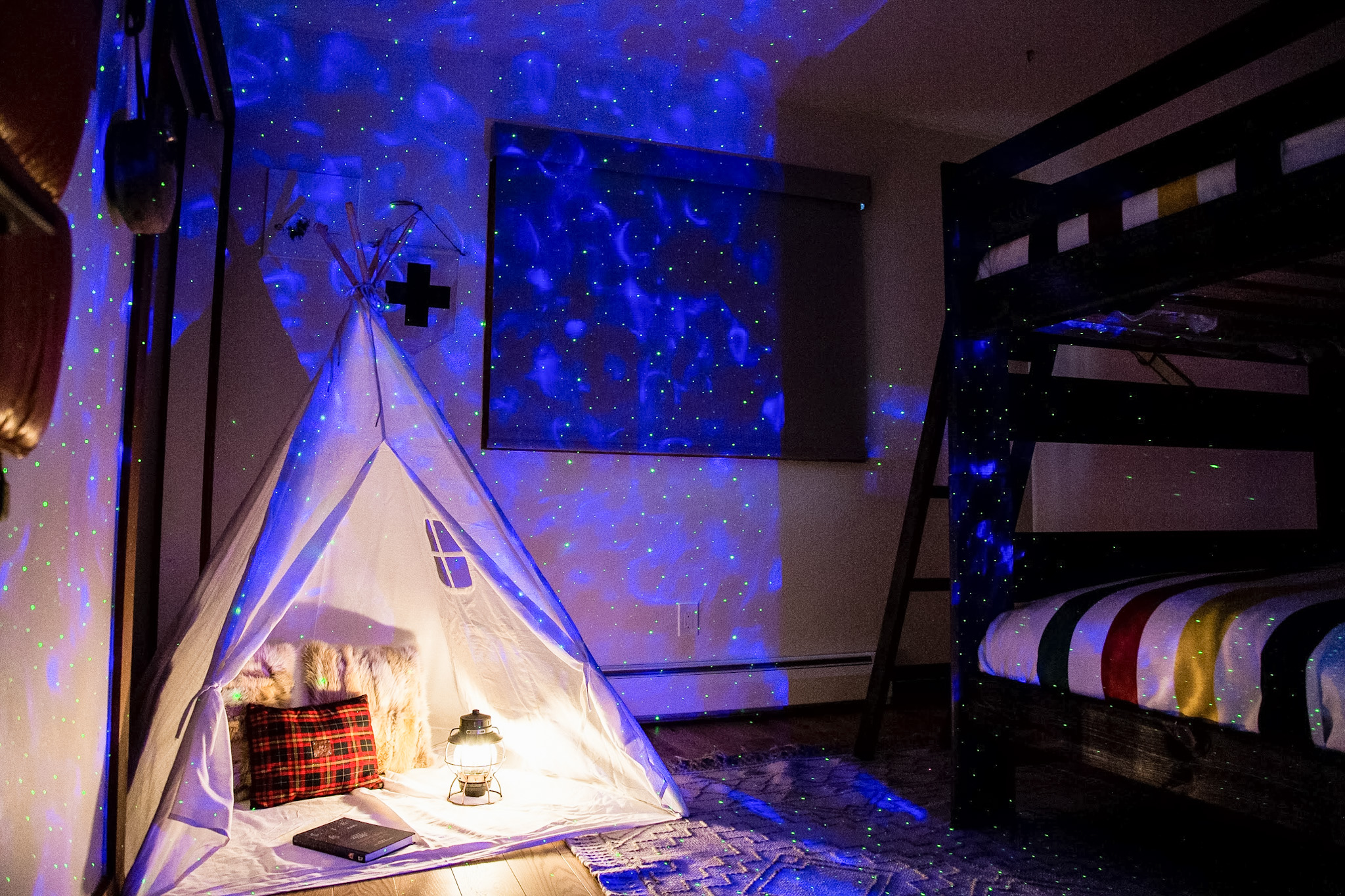 Shelf with bottles of liquor, glassware, a vase with white flowers, lemons on a cutting board, and a framed forest artwork, with stacks of books below.
