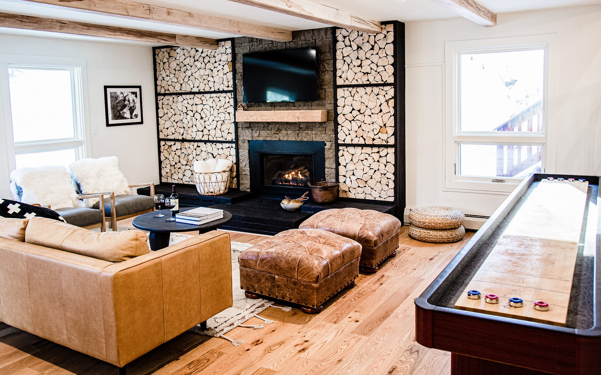 Cozy living room with a leather sofa, two tufted ottomans, two armchairs with white fur throws, a fireplace surrounded by stacked firewood, and a shuffleboard table.