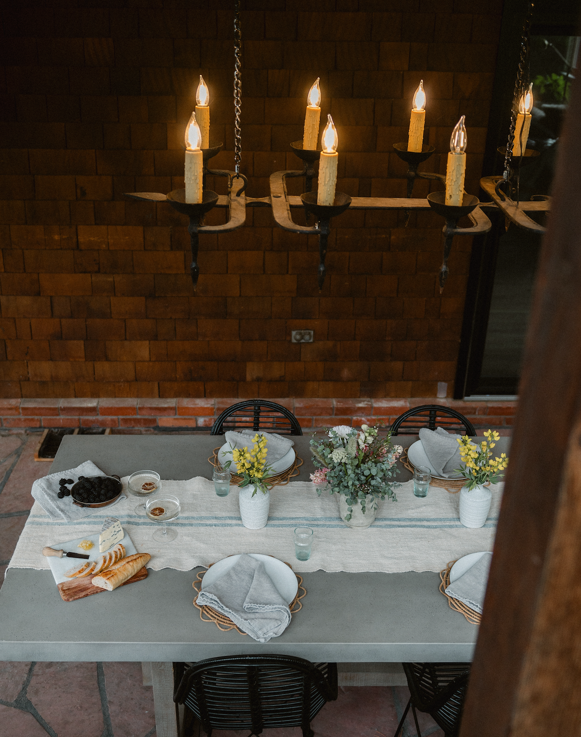 Dining table set with plates, napkins, bread, cheese, flowers, and candles hanging above, in a rustic indoor space.