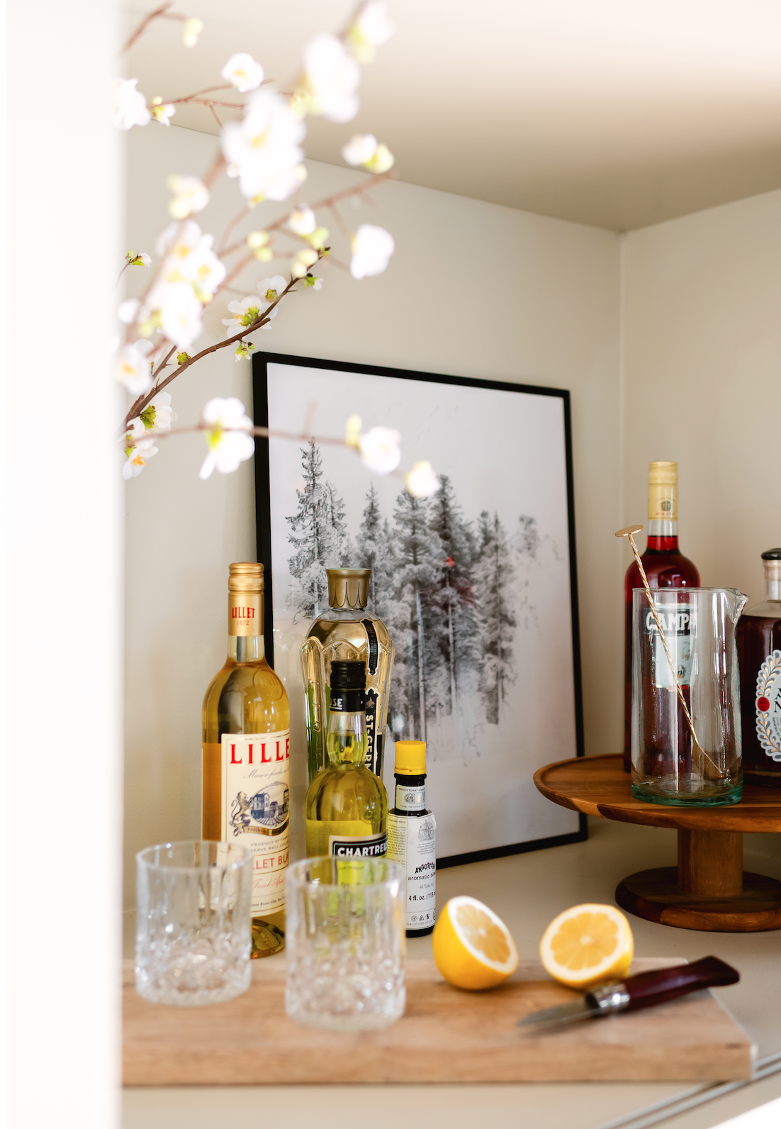 A home bar setup with bottles of Lillet, Chartreuse, Angostura bitters, two empty textured glasses, a wooden cutting board with a halved lemon and knife, and a framed black-and-white pine tree print in the background.