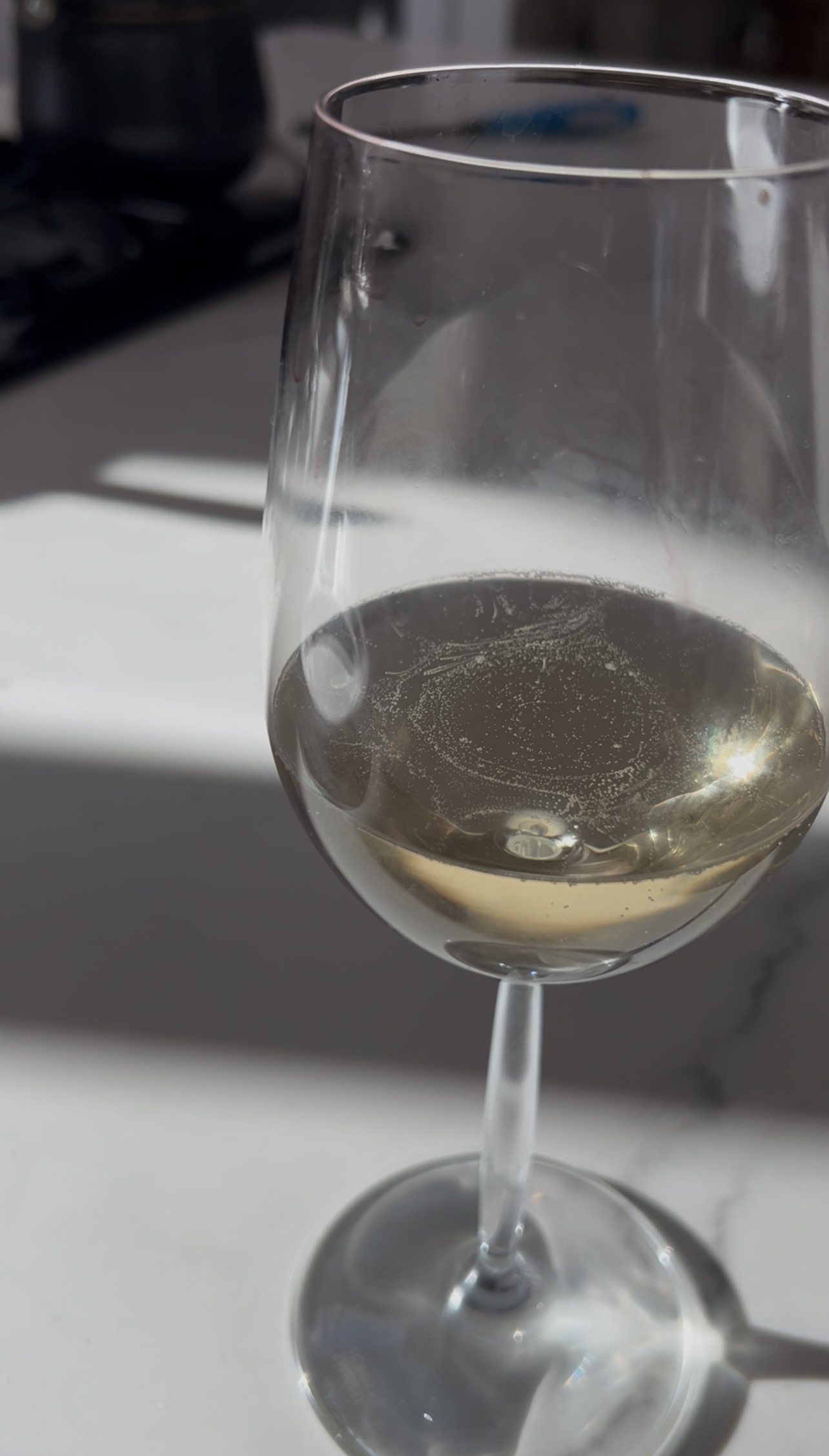 Close-up of a wine glass filled with pale yellow liquid casting a shadow on a white surface under sunlight.