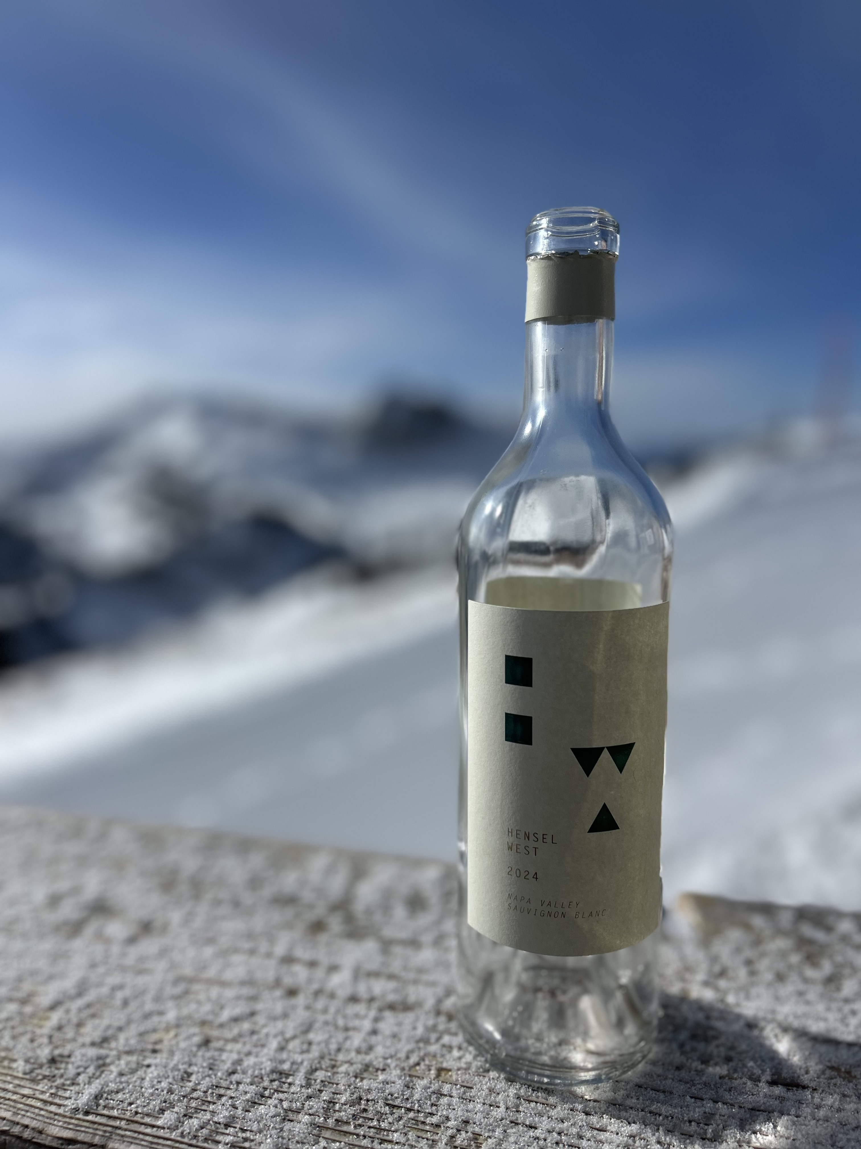 Empty Hensel West 2024 Sauvignon Blanc wine bottle on a snowy wooden surface with snow-covered mountains and blue sky in the background.