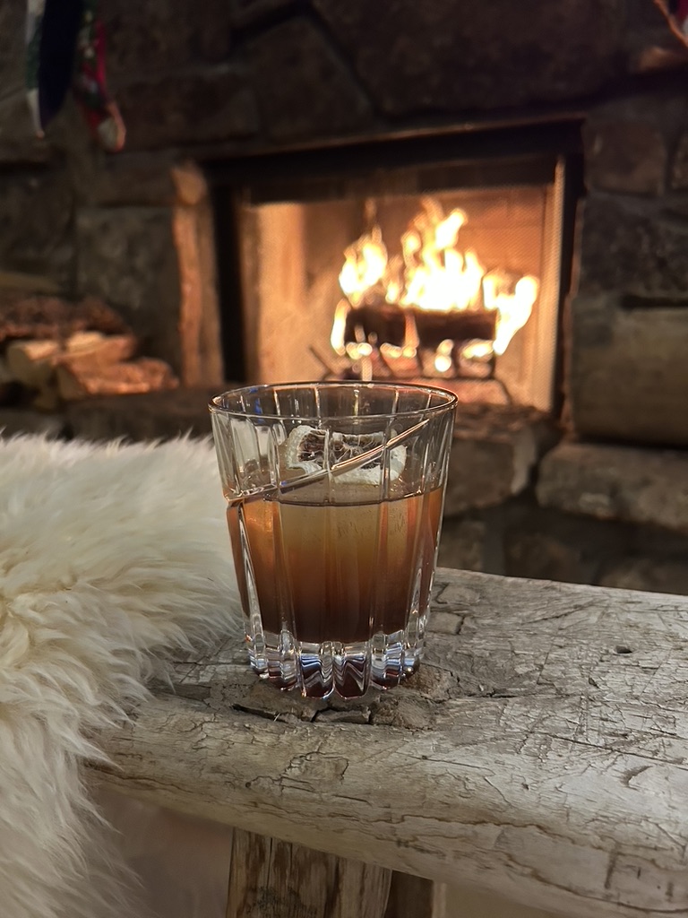 Glass of brown cocktail with a toasted marshmallow garnish on a rustic wooden bench in front of a lit stone fireplace.