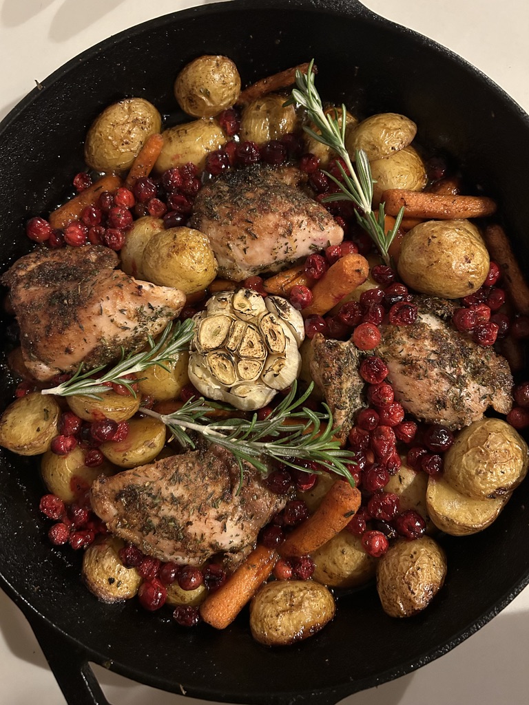Cast iron skillet with roasted chicken thighs, baby potatoes, carrots, cranberries, whole roasted garlic, and sprigs of rosemary.