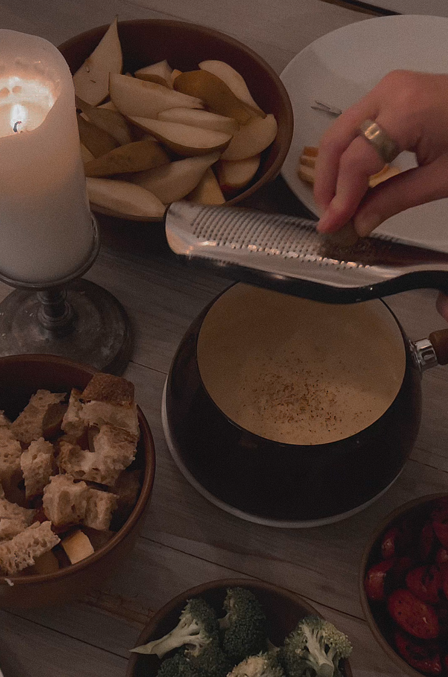 Hand grating a spice over a pot of melted cheese surrounded by bowls of bread cubes, apple slices, broccoli, and cherry tomatoes near a lit candle.