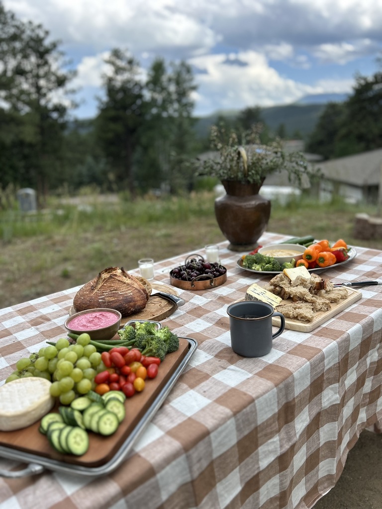 Outdoor picnic table with a checkered tablecloth holding assorted fresh vegetables, grapes, bread, cheese, cherries, and a dark mug under a cloudy sky.