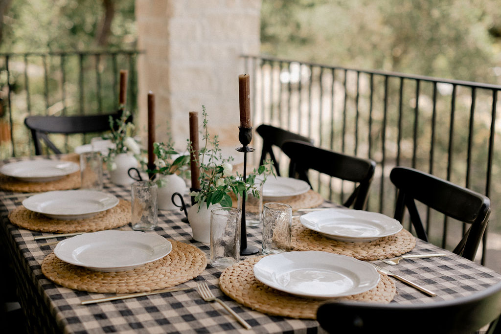 Outdoor dining table with checkered tablecloth, six place settings with white plates on woven placemats, clear glasses, brown candles, and small white vases with green plants.