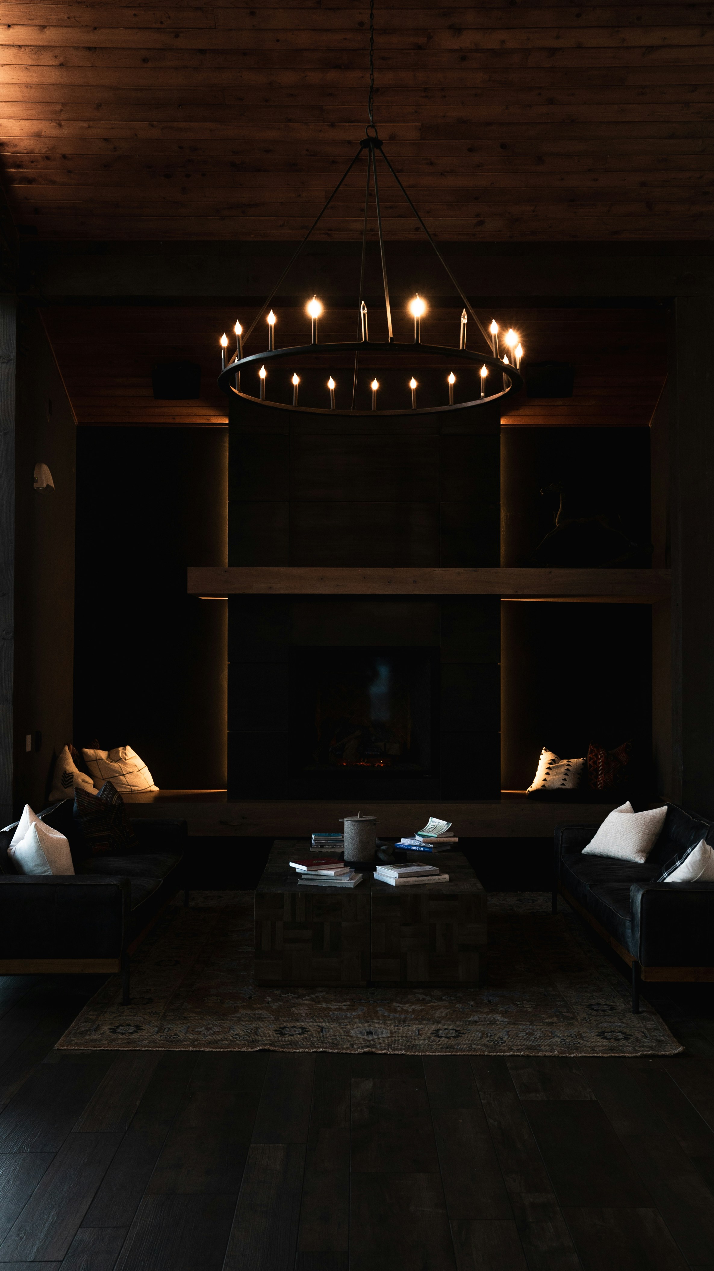 Cozy dark living room with wooden ceiling, two black sofas with white cushions, a central coffee table with books, and a glowing chandelier above.
