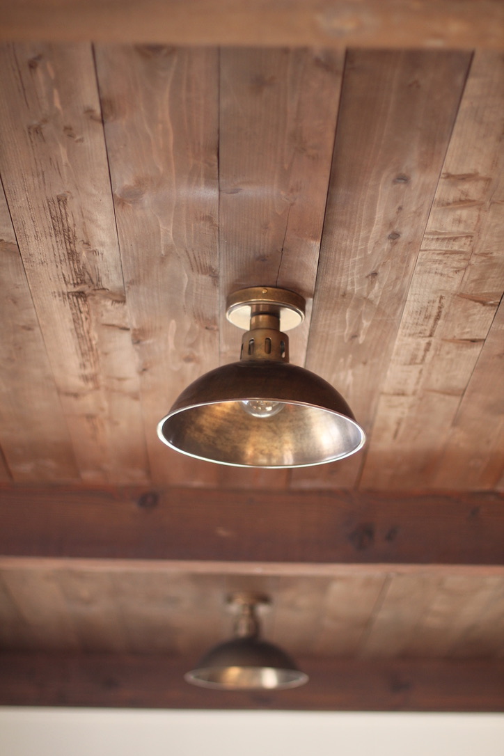 Vintage brass ceiling light fixture on a wooden plank ceiling with another similar fixture blurred in the background.