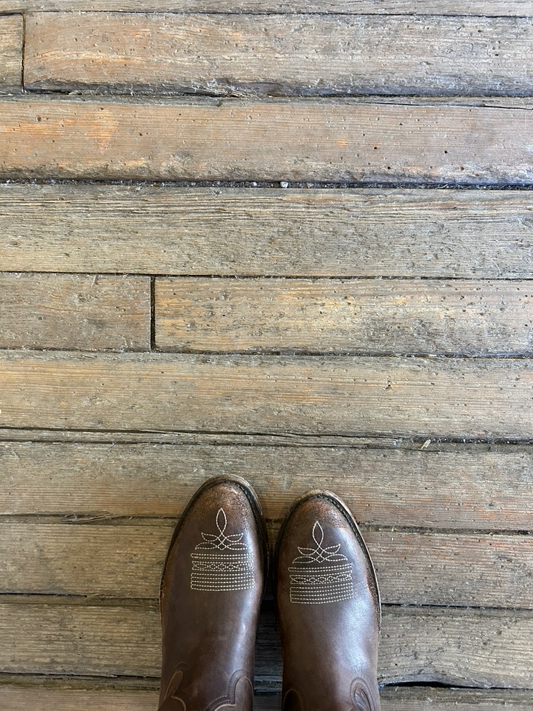 Pair of brown leather cowboy boots with stitched designs on toes standing on weathered wooden floorboards.