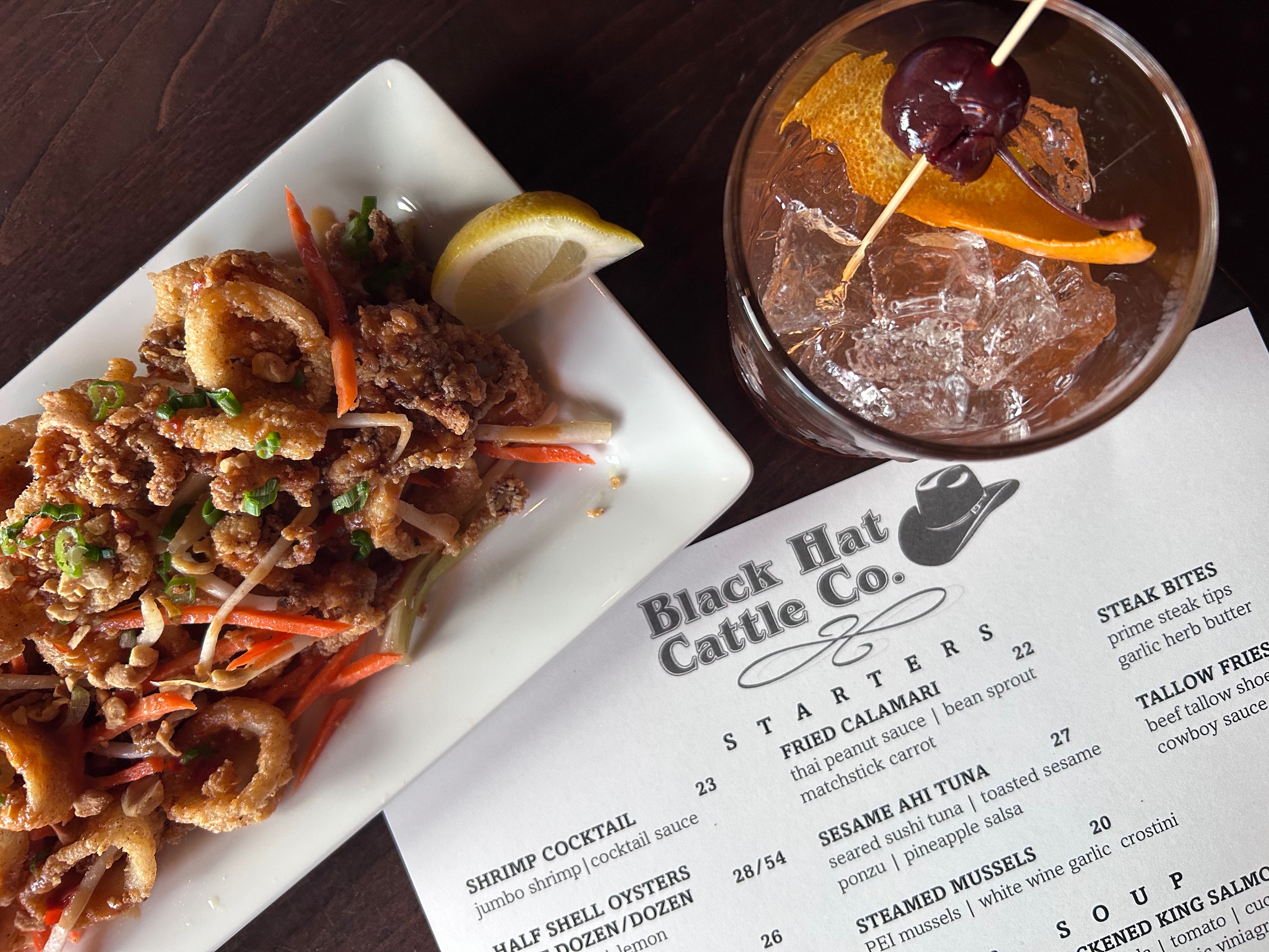 Plate of fried calamari garnished with green onions and matchstick carrots next to a n Old Fashioned cocktail with an orange peel and cherry on a skewer on a Black Hat Cattle Co. menu.