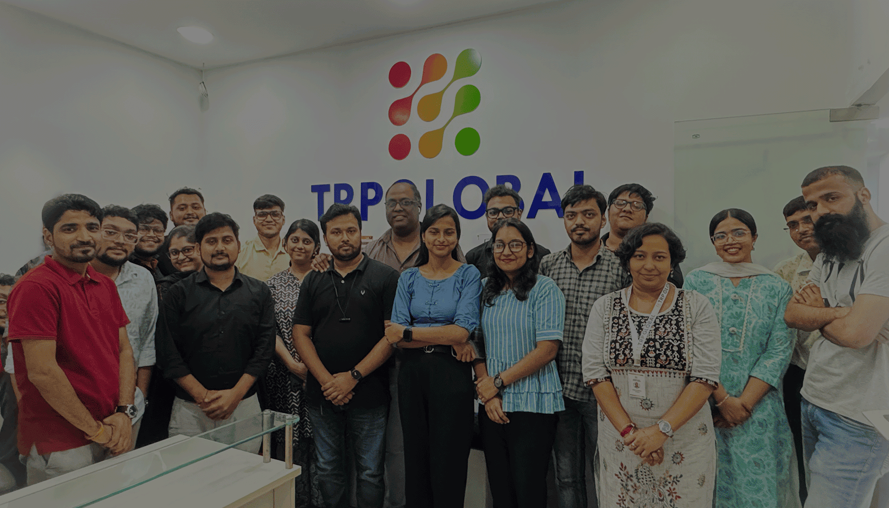 Group of diverse colleagues standing together inside an office with a colorful logo and TRPGLOBAL text on the wall behind them.