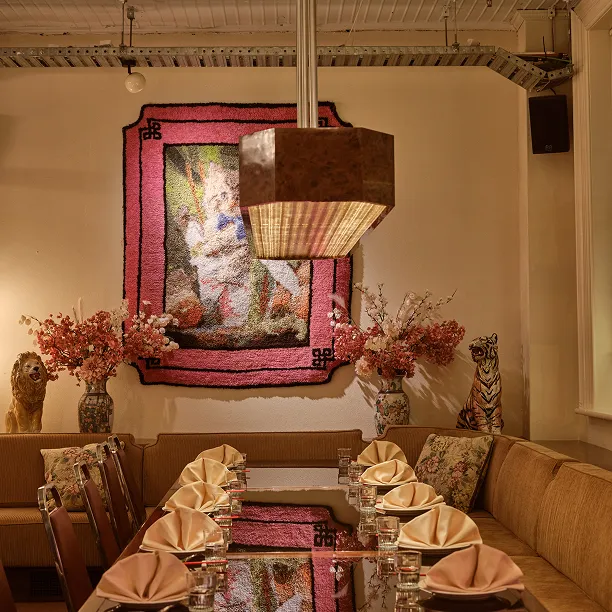Cozy dining area with a long table set with folded napkins and glasses, floral cushions on beige seating, a hanging light fixture, and decorative lion and tiger statues with flower vases under a colorful textile artwork on the wall.