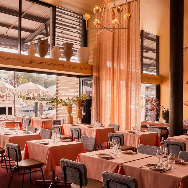 Elegant restaurant interior with tables covered in peach tablecloths, gray chairs, decorative pottery on a high shelf, and large windows overlooking outdoor umbrellas.