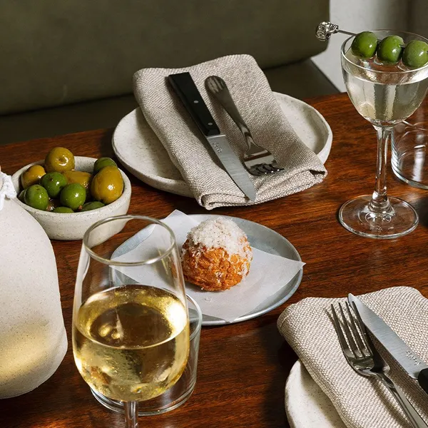 Table set with white wine in a glass, a bowl of green and yellow olives, a small plate with a breaded appetizer topped with salt, and two plates with beige napkins holding a knife and fork each, plus a cocktail glass garnished with three green olives.