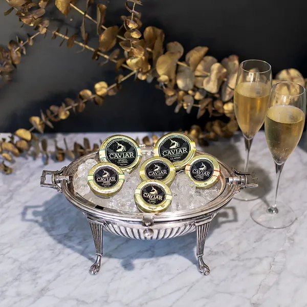 Six tins of Aquatir Classic Caviar on ice in a metal serving dish with two glasses of champagne on a marble table.