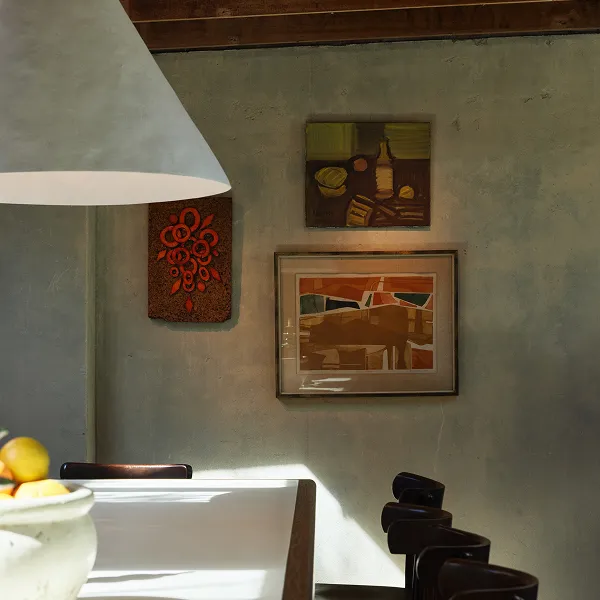 Dimly lit dining area with a hanging lamp, three framed abstract paintings on a textured wall, and part of a white table with dark chairs.