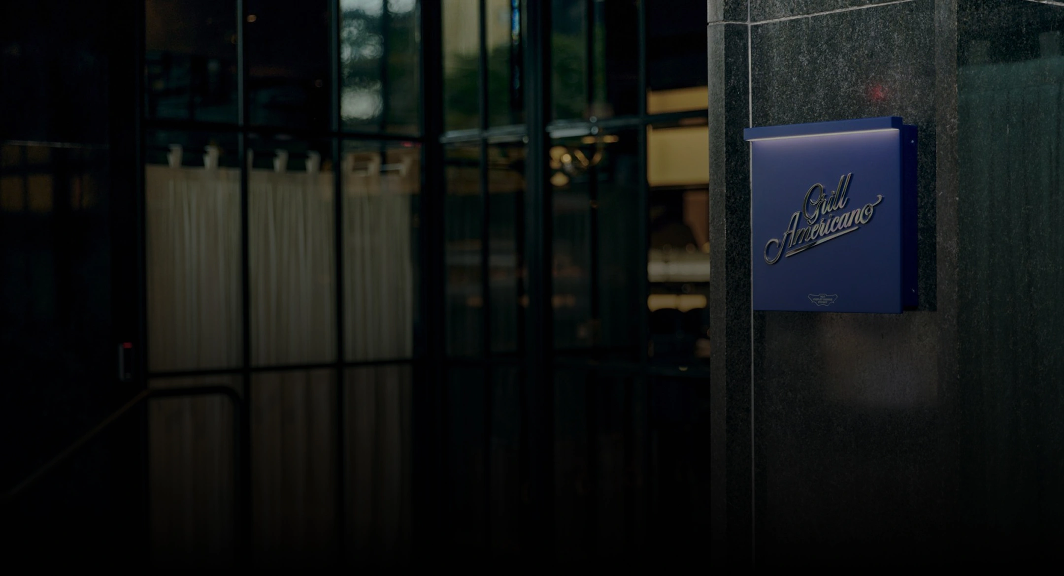 Blue illuminated sign with cursive text 'Grill Americano' mounted on a dark stone wall.