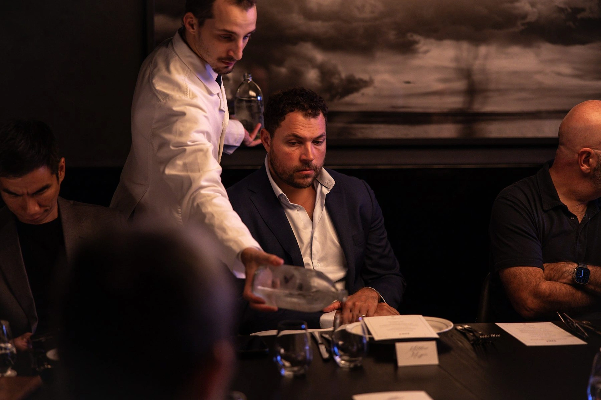 Waiter in white jacket pours water into glass for man in dark suit at dimly lit dinner table.
