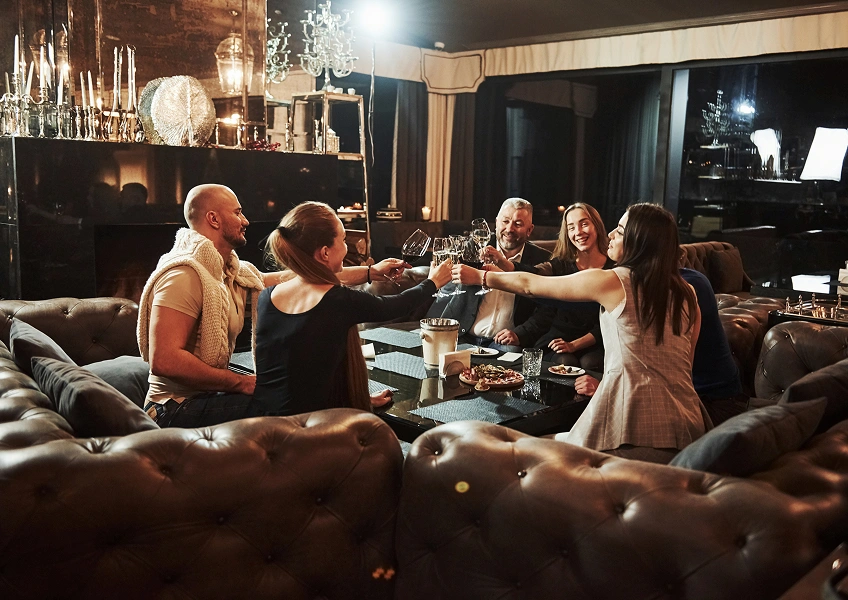 Group of friends sitting on sofas around a table, toasting drinks in a cozy living room.