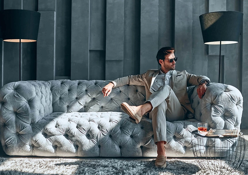 Man in beige suit and sunglasses sitting on a tufted gray sofa between two standing lamps.