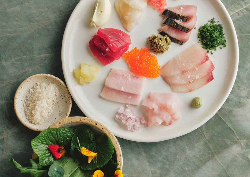 Assorted sashimi and garnishes including fish slices, roe, herbs, and salt on a white plate.